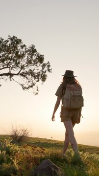 Young woman in safari outfit and hat walks along mountain trail at sunset with backpack. Golden rays create dreamy atmosphere of freedom, adventure, healthy lifestyle and stylish exploration in slow