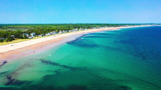 Drone flying above Saco, Maine coastline shows people enjoying peak-season beach life. Sand full of tourists, vacation homes nearby, waves crash on shore. A beautiful summer day on the Atlantic.