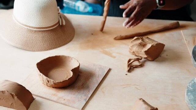 Hands roll a clay coil and shape a rustic bowl using hand-building pottery techniques on a sunlit table, with a straw hat nearby.