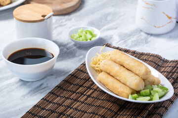 A serving of fried pempek lenjer, traditional Indonesian fishcake, arranged with noodles and diced cucumber. A bowl of cuko sauce is placed beside for dipping