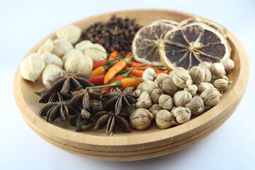 Cardamom, cloves, star anise, dried lemon, candlenut, and chili peppers arranged in a wooden bowl on white background