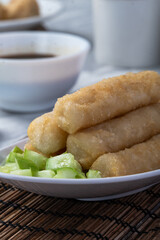 A serving of fried pempek lenjer, traditional Indonesian fishcake, arranged with noodles and diced cucumber. A bowl of cuko sauce is placed beside for dipping