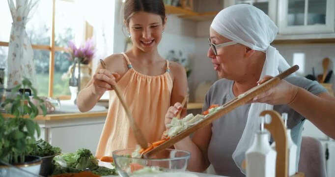 A smiling mother with a scarf on her head due to chemotherapy makes a vegetable salad in the kitchen with her cheerful daughter