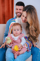 Smiling parents on sofa entertain their baby daughter in pink overalls with yellow duck and green...