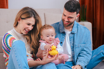 Smiling parents on living room sofa entertain their baby daughter in pink overalls with yellow duck...