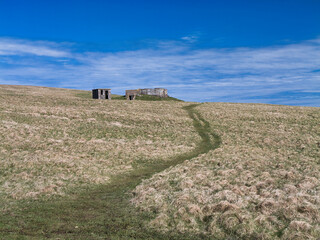 A remote coastal path leads past historic World War II buildings under a clear blue sky in Skaw, Unst, Shetland.
