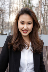 A smiling young woman with braces and wavy hair poses outdoors in a black blazer and white shirt