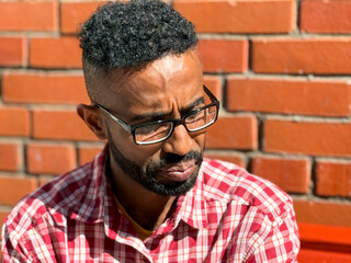 Portrait of a young man of African-American nationality wearing glasses with a beard sitting on a bench. A black guy in a plaid shirt on the background of a brick wall. Portrait of a black man. 