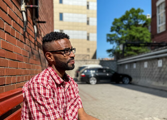 Portrait of a young man of African-American nationality wearing glasses with a beard sitting on a bench. A black guy in a plaid shirt on the background of a brick wall. Portrait of a black man. 