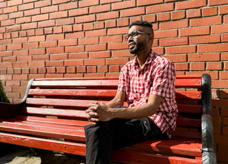 Portrait of a young man of African-American nationality wearing glasses with a beard sitting on a bench. A black guy in a plaid shirt on the background of a brick wall. Portrait of a black man. 