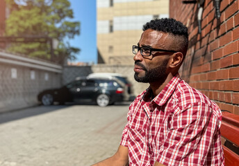 Portrait of a young man of African-American nationality wearing glasses with a beard sitting on a bench. A black guy in a plaid shirt on the background of a brick wall. Portrait of a black man. 