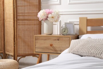 Bedside table with alarm clock and flowers near bed indoors