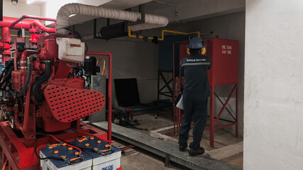 A worker in a dark room inspects a red fire pump system with pipes and a control panel, near a red cabinet.