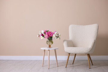 Comfortable armchair and table with vase of flowers near beige wall indoors, space for text