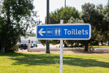 A blue sign with a spelling error for “toilets” points left to public restrooms in a park area in Lisbon, Portugal.