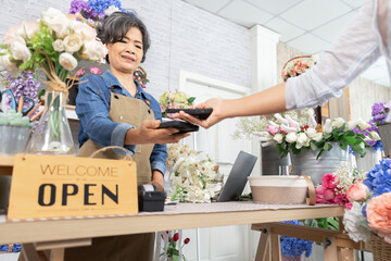 Asian senior woman, florist, small business, owner, entrepreneur handling electronic payment from customer at cozy flower shop, showing cashless service, trust, lifestyle, technology, age 60s retired