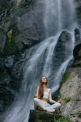 Woman meditating near waterfall in white activewear surrounded by rocks and green moss, showing...