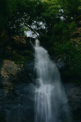 Majestic waterfall cascading down rocky cliffs surrounded by dense green forest foliage under a...