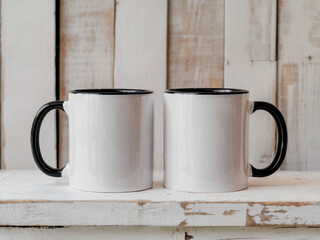 A product photography shot of two white ceramic coffee mugs with black handles and black interior rims, positioned side by side on a distressed white wooden shelf
