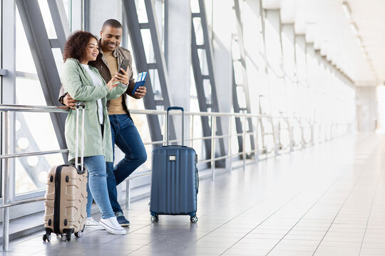 Young Black couple at airport looking at smartphone together, checking flight details or travel plans, luggage by side, casual and connected modern travelers, copy space