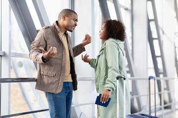 Young Black couple arguing in airport, having intense conversation, upset expressions, holding...