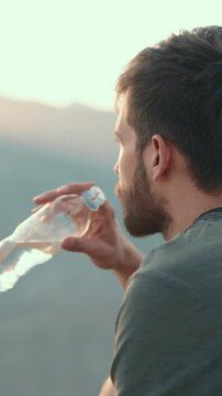 Closeup of male hiker sitting on mountain peak at sunrise, thoughtfully drinking pure water from plastic bottle. Inspiring moment of freedom, healthy lifestyle, mindfulness and adventure in scenic