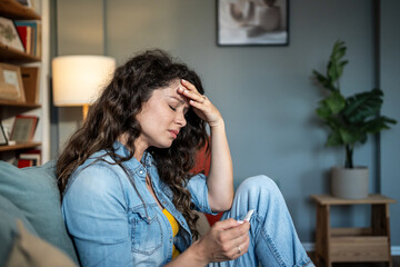 Sad woman holding pregnancy test sitting on sofa at home