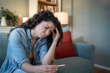 Sad woman holding pregnancy test sitting on sofa at home