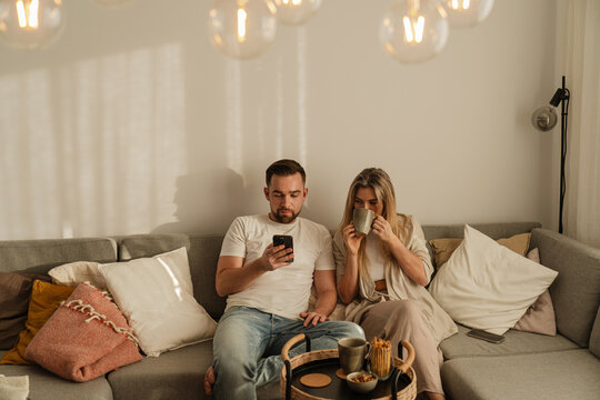 Happy Couple Sitting Together on Sofa Using Smartphone and Drinking Coffee