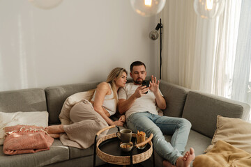 Couple Relaxing on Sofa at Home Using Smartphones with Snacks Nearby