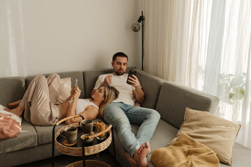 Couple Relaxing on Sofa at Home Using Smartphones with Snacks Nearby © blackday