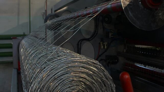 A close-up view of a large, cylindrical roll of galvanized wire mesh, likely a fixed-knot fence, mounted on a piece of industrial machinery.