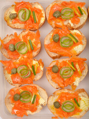 An overhead view shows eight canapes with a creamy spread topped with vibrant orange carrot prepared as vegan salmon, green leeks, and capers, arranged in a clear plastic container