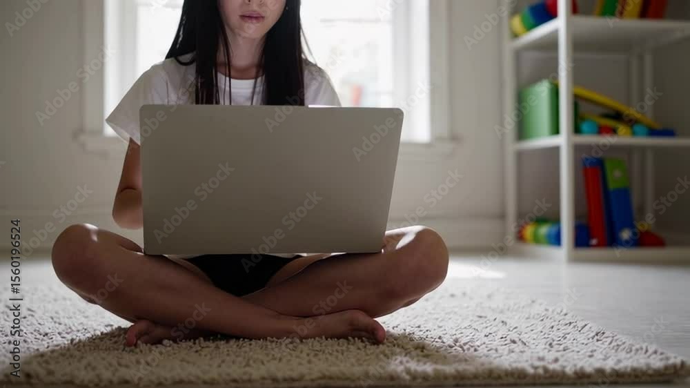 Poster comfortable pupil sitting cross legged on a carpet, using a laptop in their bedroom for online educa