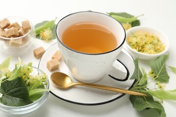 Tasty linden tea in cup, spoon, sugar cubes, leaves and flowers on white table, closeup