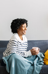 Happy young Latin American woman enjoying hot drink on sofa on a cozy winter day at home