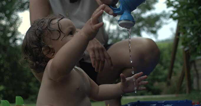 Baby raising arms playfully as water pours from a plastic watering can in slow motion, joyful outdoor moment with parent in background and garden surroundings