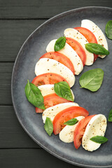 Italian Caprese salad with cherry tomatoes, mozzarella, basil on dark plate on black wooden background. Vertical photo