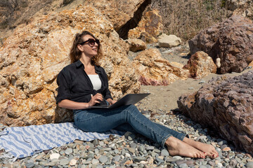Woman Laptop Beach, works outside near rock formation on sunny day for remote job.