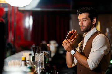 A handsome and focused professional bartender concentrating as he prepares a craft cocktail with a copper shaker in a stylish, dark bar at night.
