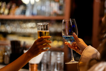 Two female friends raising glasses of beer and a blue cocktail for a toast in a stylish bar. Concept of celebration, friendship, and happy hour on a night out in Bangkok.