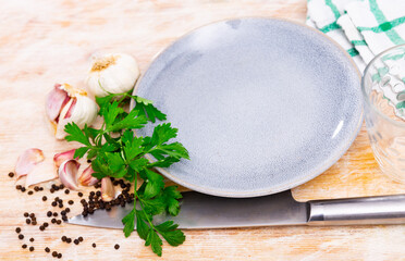 There is knife, garlic, fresh parsley and corn of black pepper next to white plate. Fragrant ingredients for cooking homemade dishes.