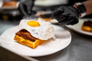 Close-up of a chef's gloved hands plating a savory waffle dish with fried egg, pulled meat, and melted cheese on a white plate, showcasing gourmet presentation and texture.