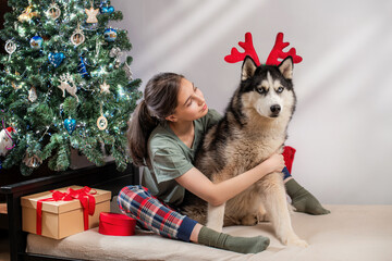 Young woman with brown hair hugging a husky dog with red reindeer antlers next to a decorated Christmas tree. New Year celebration at home with a pet