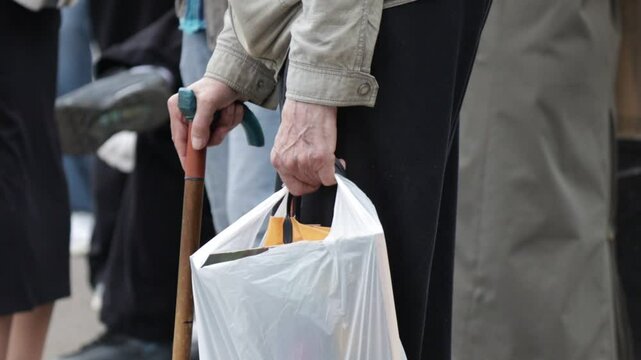 Elderly man rests on cane with one hand, in second hand he holds bag. Man is surrounded by other people