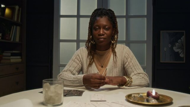 Zoom in of African American tarot reader sitting at round table with cards, plates and glass of minerals and crystals. She looking straight to camera and smiling