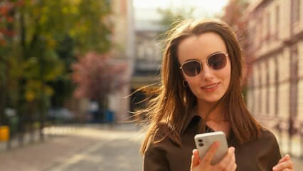 Happy smiling laughing beautiful stylish business woman businesswoman girl female employer using mobile phone smartphone app network scrolling social media walking in city outdoors gadget addict sms