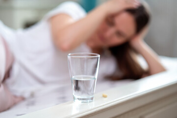 Glass of water with a pill in the background, sick woman holding her head showing signs of headache...