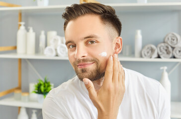 Skin care for men. Man enjoying skin care routine applying moisturizer with smile in bathroom.
