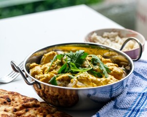 A serving of chicken korma, a creamy dish with tender pieces of chicken in a rich, golden-yellow sauce, garnished with fresh arugula leaves. It is presented in a metal bowl